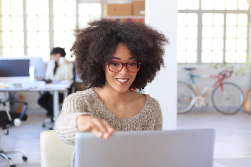 Smiling young woman with red eyeglasses finishing her work at the office and closing laptop. Looking...