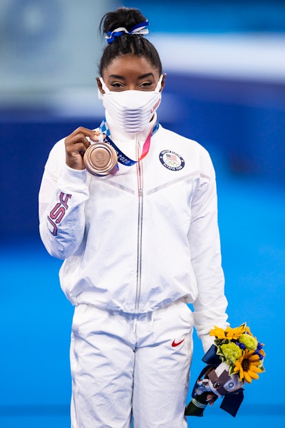 Simone Biles, wearing an all-white Team USA tracksuit, shows off her bronze medal at the 2021 Olympi...