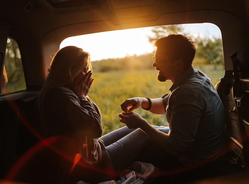 Romantic young couple sitting in open car trunk, enjoying sunset in spring nature while taking a bre...