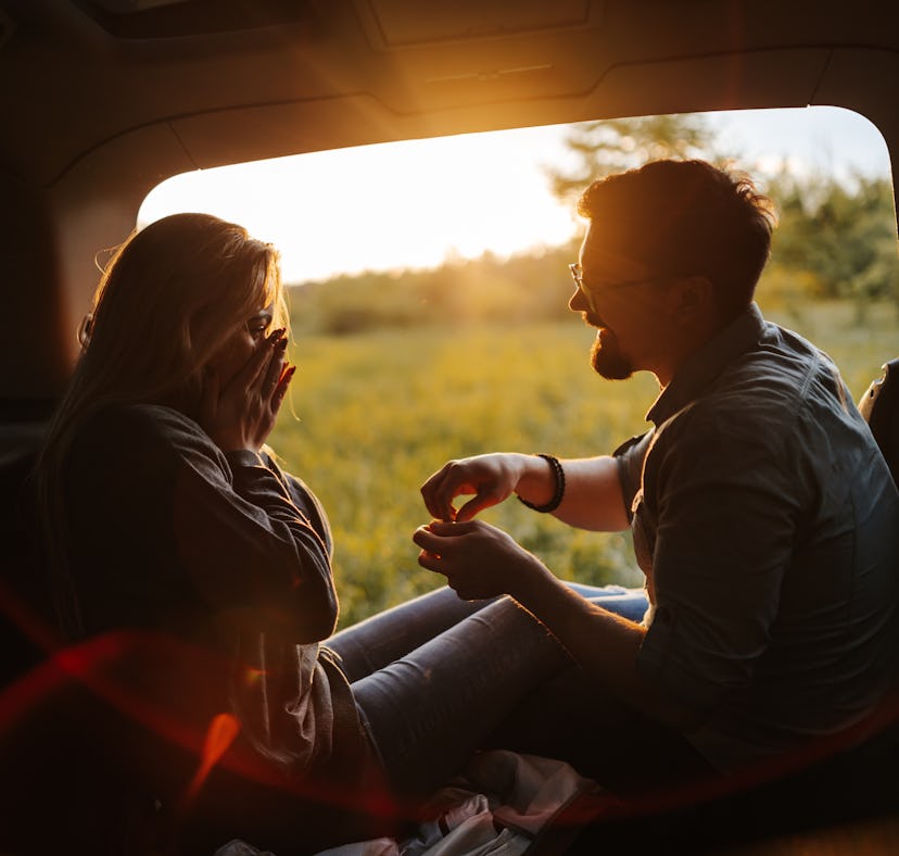 Romantic young couple sitting in open car trunk, enjoying sunset in spring nature while taking a bre...