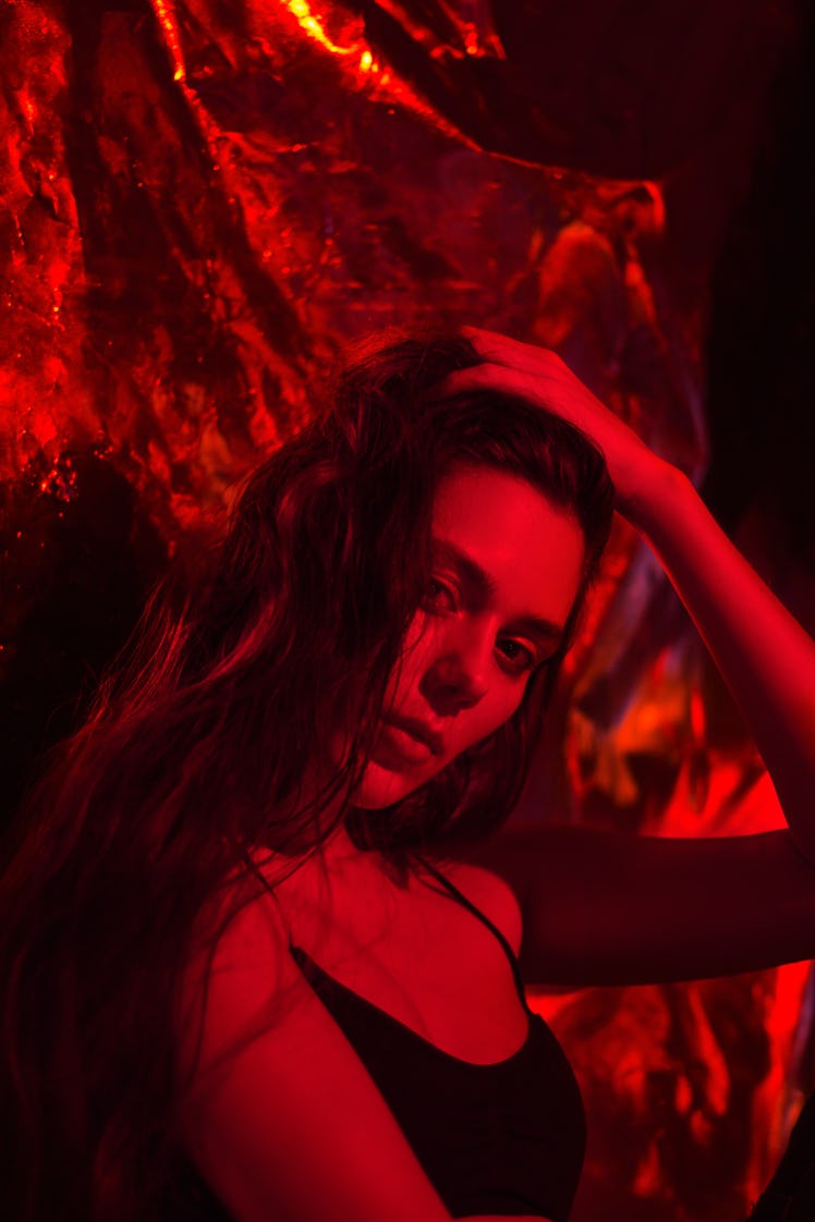 woman lit by red light, running fingers through hair as she peers into camera and reflects on the me...
