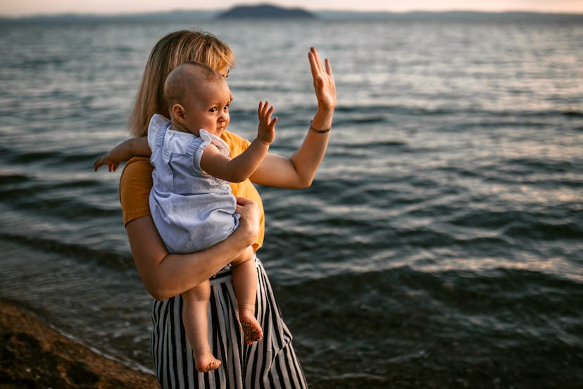 Mother and baby girl enjoy coastal walk and beautiful sunset on the sandy beach, waving to the boats