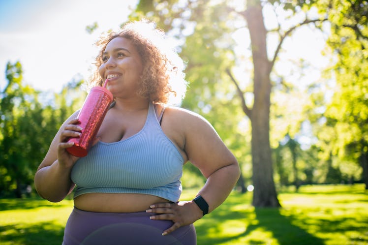 Young woman in purple workout attire stands in park drinking from a red reusable water bottle