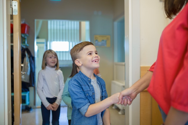 A young child entering a classroom, and shaking hands with an adult.