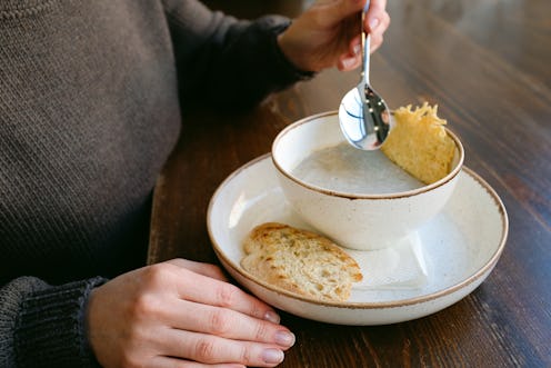 A young woman or girl eats vegetable or mushroom soup puree from a white stylish plate, at a wooden ...