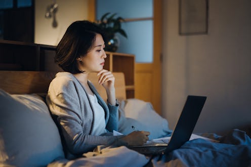 Busy young Asian woman sitting on bed, working from home using laptop and handling paperworks till l...