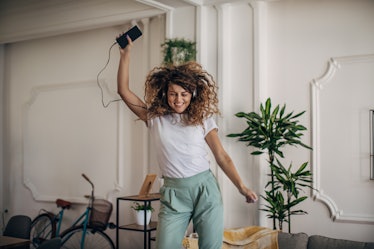 One woman, beautiful woman standing in living room at home. She is using smart phone, dancing and li...