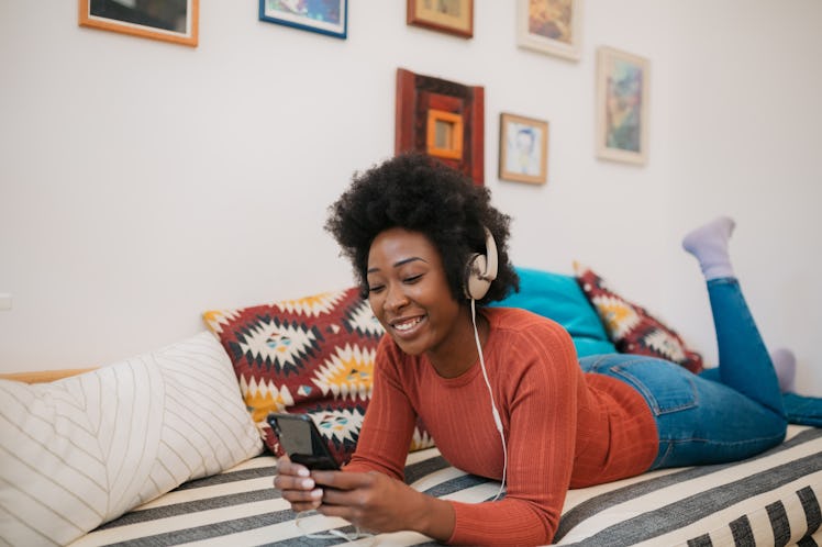Young cheerful African American woman relaxing at home, using a smart phone to listen to music and s...