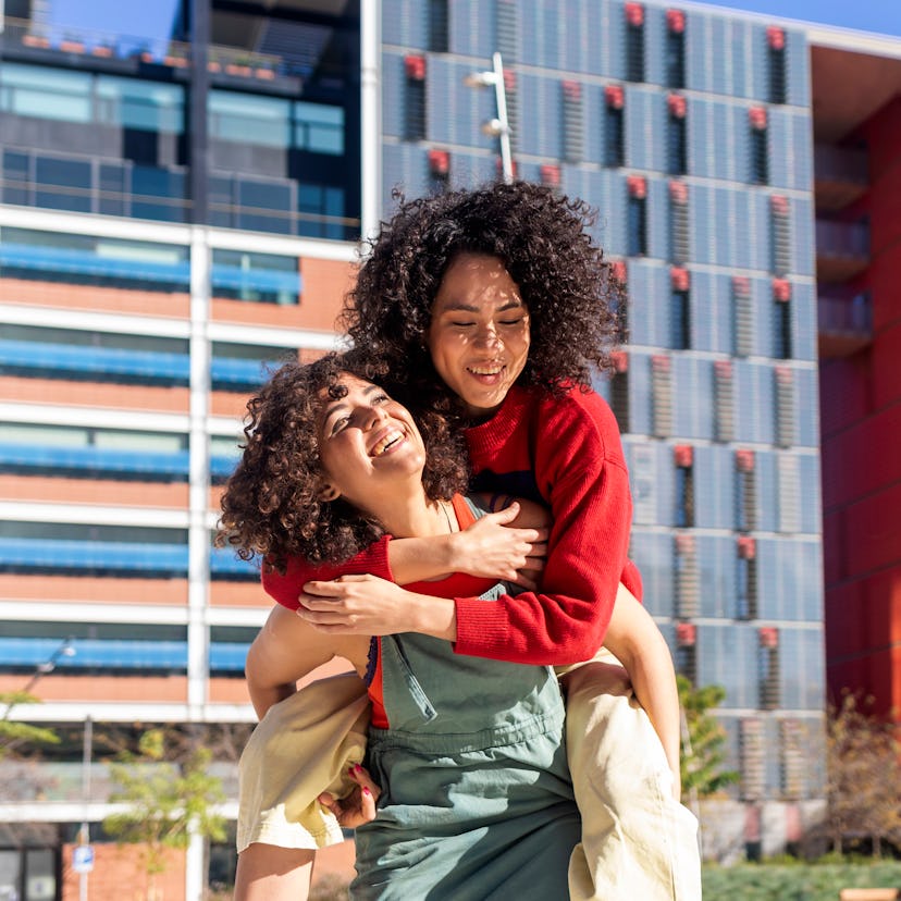 two female friends smiling as one gives the other a piggy back ride as they chat about leo libra fri...