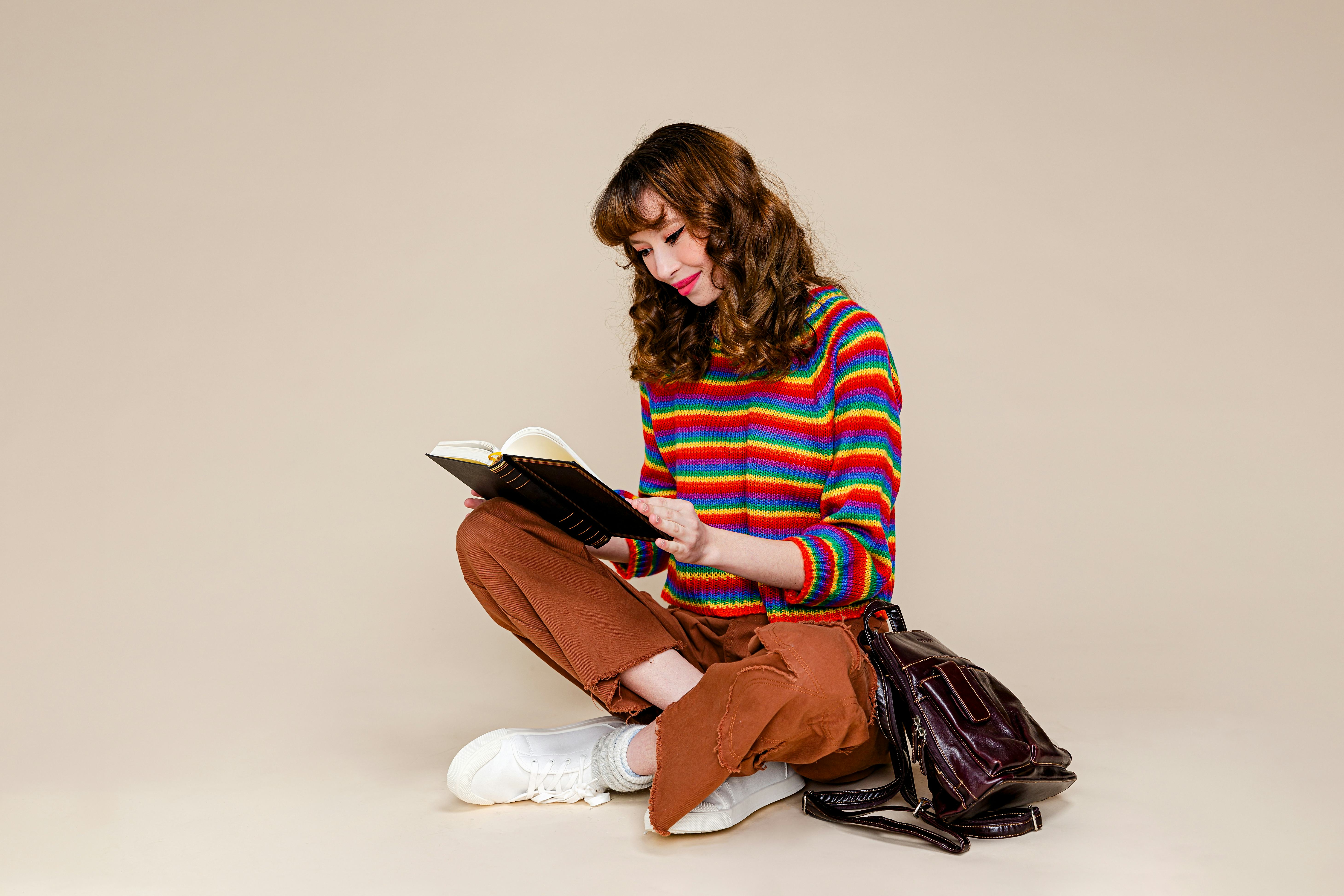 A portrait of a happy young woman sitting on a floor reading a book with her backpack in front of pl...