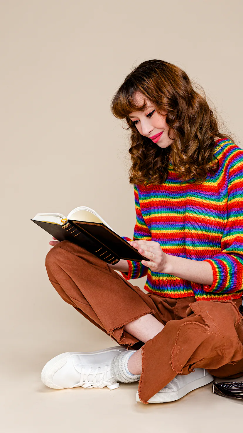 A portrait of a happy young woman sitting on a floor reading a book with her backpack in front of pl...
