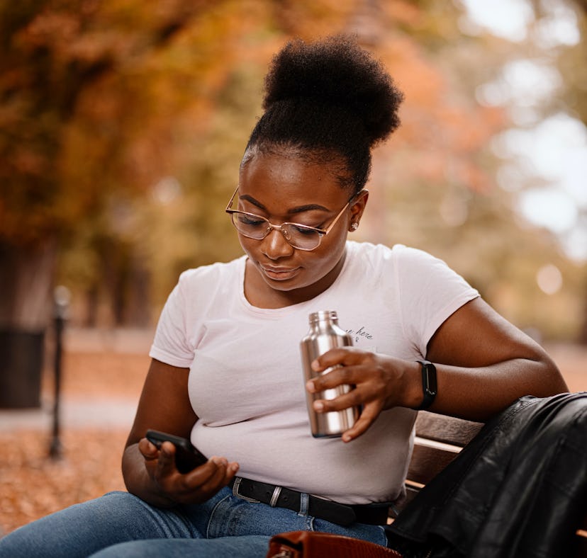 Young black woman uses the smartphone while sitting on a park bench. Autumn concept