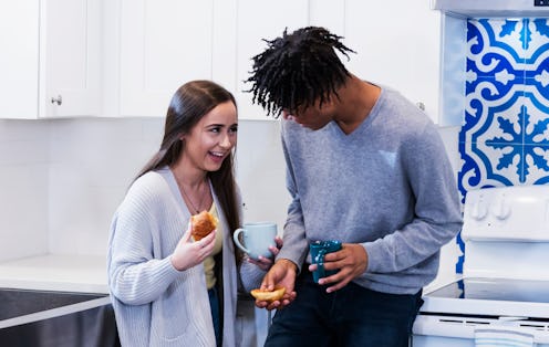 A young multiracial couple standing in the kitchen conversing, eating breakfast and drinking coffee....