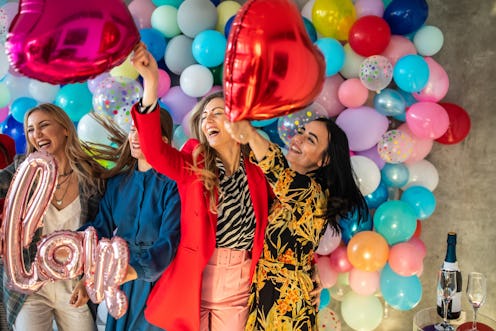 Happy female friends standing in front of wall with bunch of balloons and having fun with helium bal...