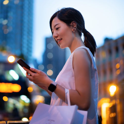 Young Asian woman using smartphone while walking down in city street against illuminated street ligh...