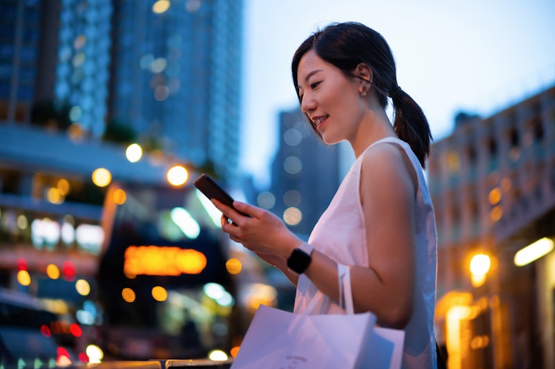 Young Asian woman using smartphone while walking down in city street against illuminated street ligh...