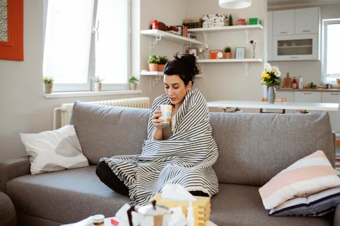 Sick woman covered with a blanket sitting in bed with high fever and flu. Pills and glass of water o...