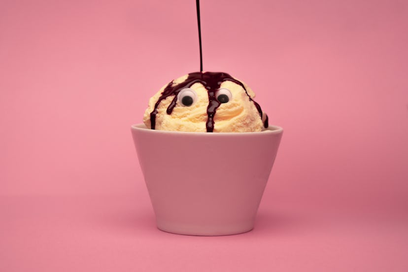 Ice cream with a funny face in a small bowl, as chocolate topping is poured on their head.