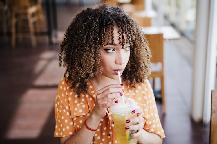 A woman drinking a hard kombucha drink at a restaurant thinks of Instagram captions for her drink.