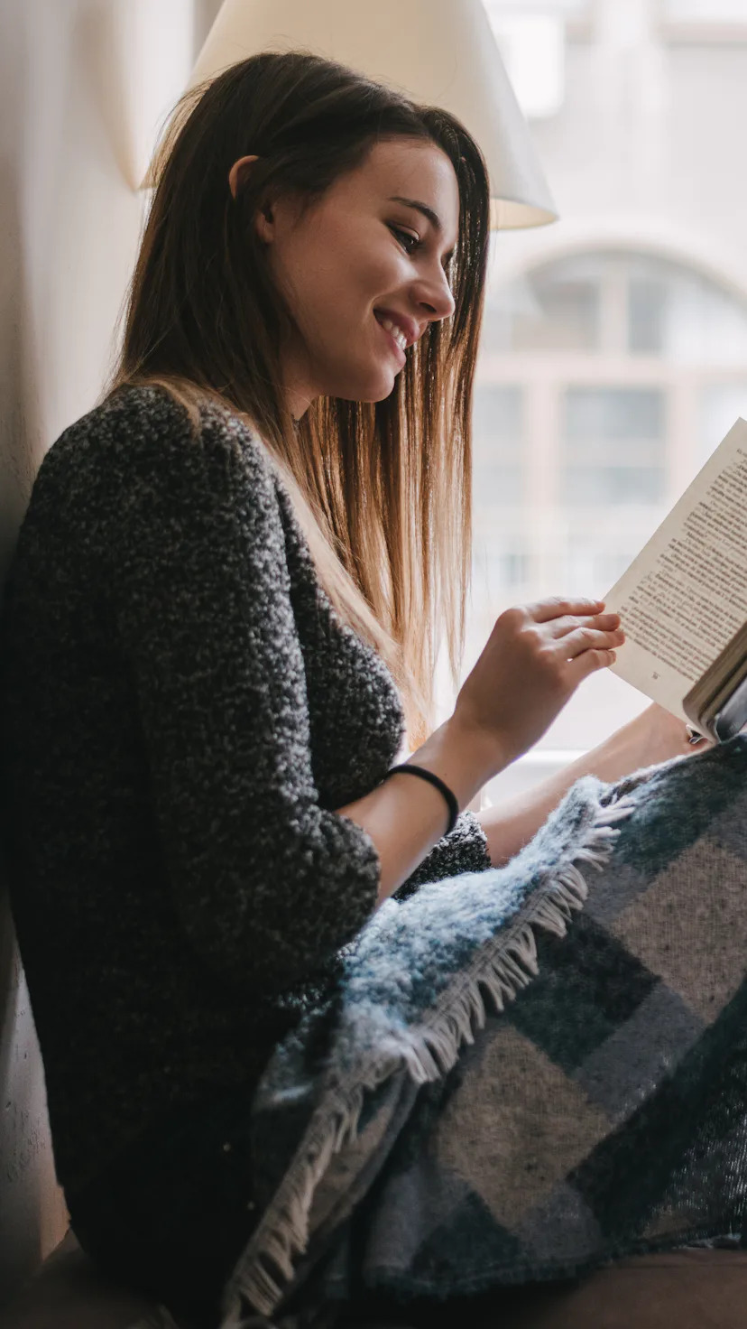 Pretty young woman reading a book near window in appartment