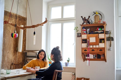 Female friends sitting at table in kitchen and talking. Women chatting at home with coffee cup on ta...