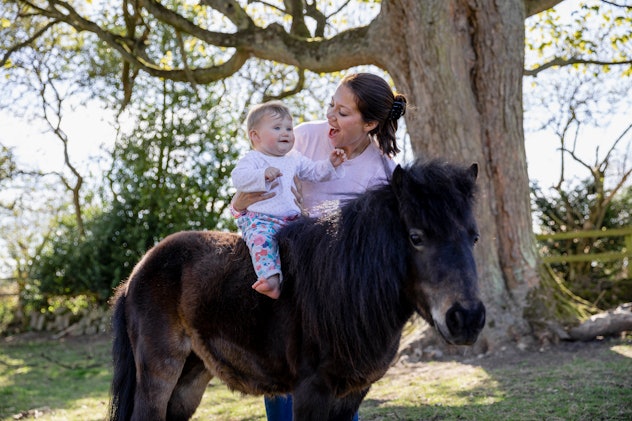 A baby girl sitting on a pony in a field while being supported by her mother who is standing next to...