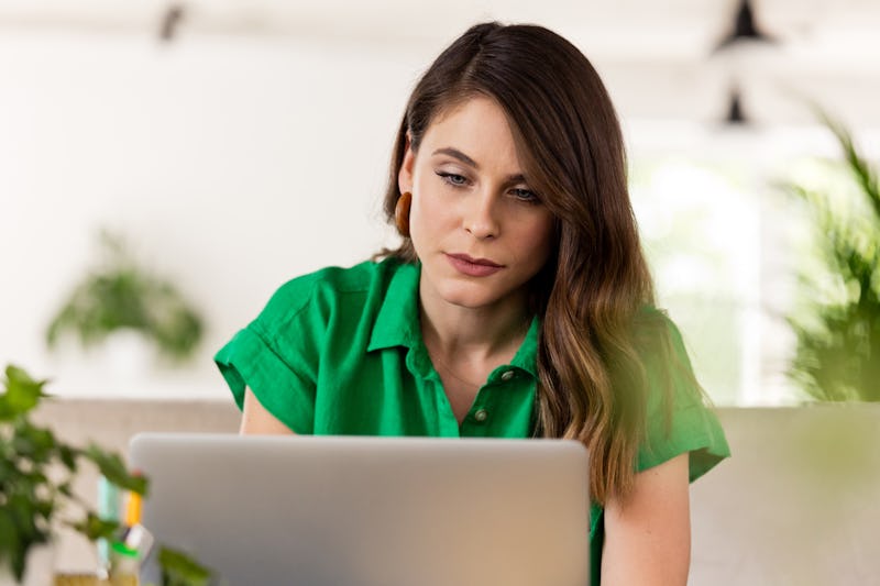 Thoughtful young woman wearing green clothes sitting on sofa in the creative workplace and using lap...