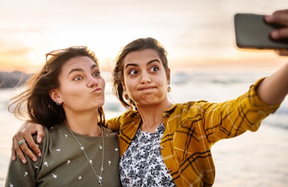 Two young female friends making funny faces while taking selfies together on a beach at sunset in th...