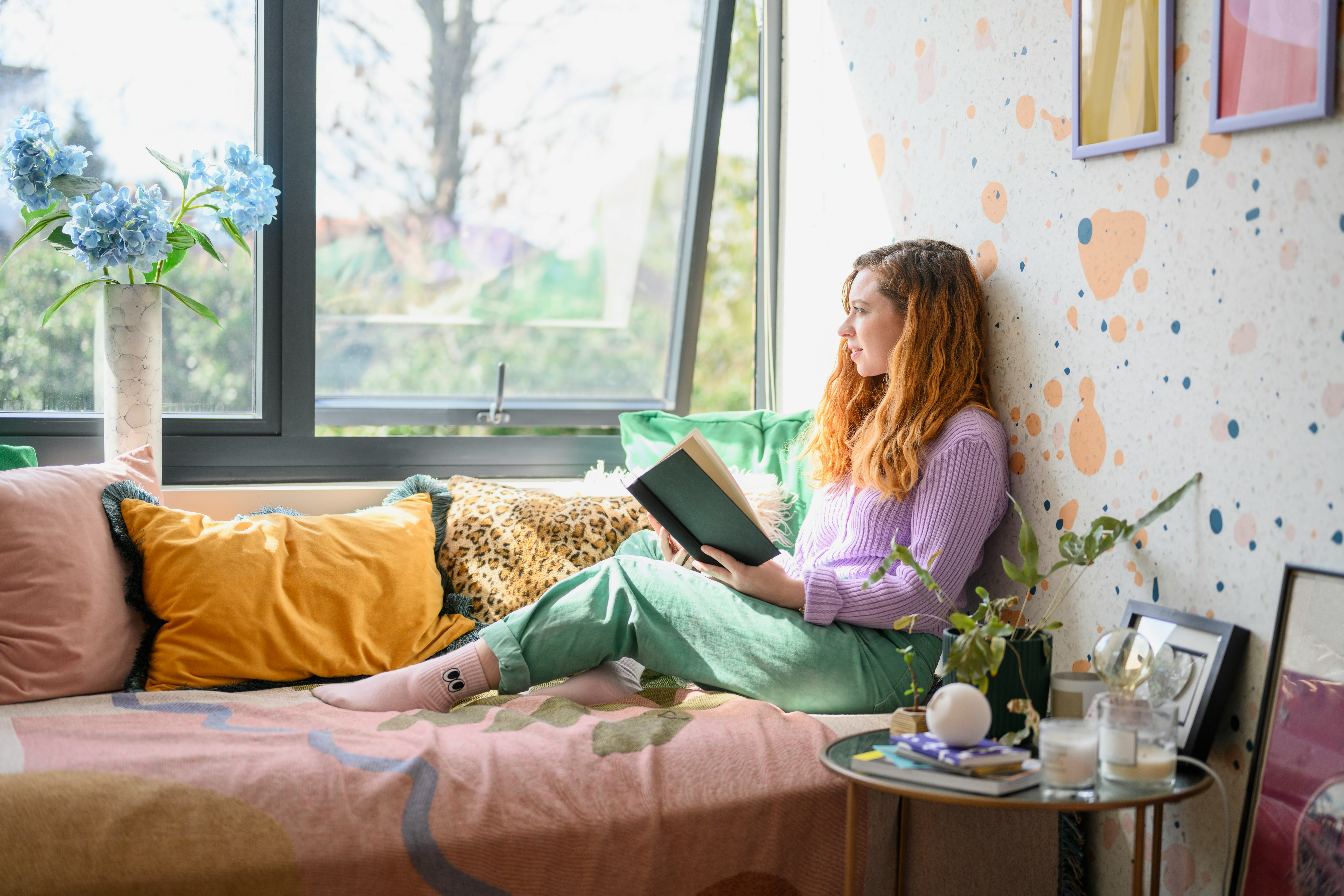 Full length side view of casually dressed redhead in mid 30s reclining on sofa and pausing from her ...