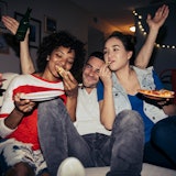 Two women and one man sitting on a couch eating pizza and partying.