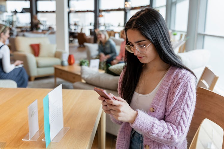 A young woman sitting at a dining table in a furniture store while using her mobile phone. There are...