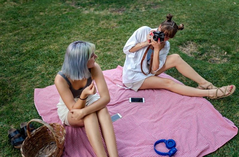 Two women take photos of each other on a picnic blanket, drifting apart as friends.