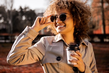 Beautiful young woman smiling wearing sunglasses and drinking a takeaway cup of coffee outside