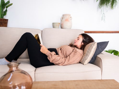 Side view full body of concentrated female reading book while lying on comfortable couch in modern l...