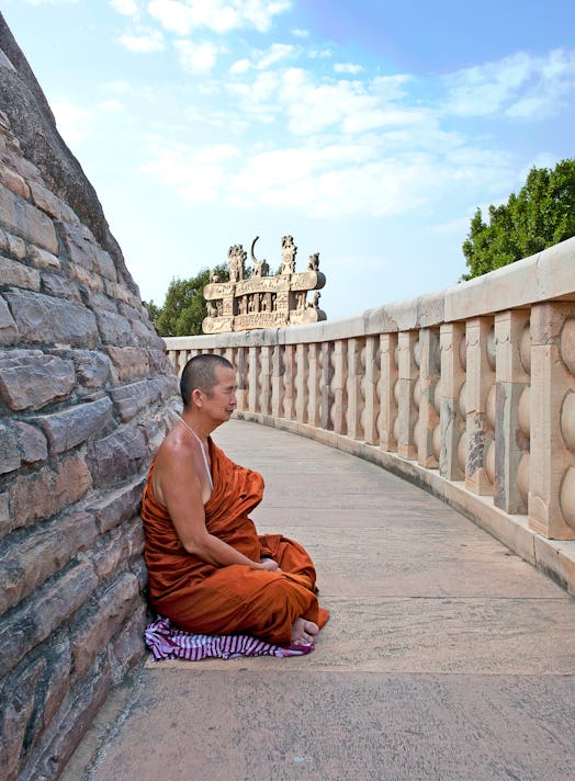 Buddhist monk in traditional robes sits in contemplation next to a Stupa at Sanchi, Madhya Pradesh, ...