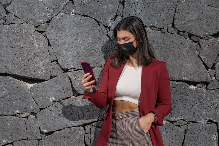 Young female wearing stylish outfit and mask browsing mobile phone near stone wall with hand in pock...