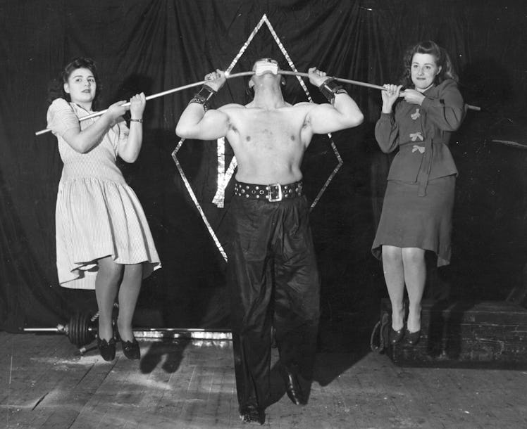 1962:  Alexander Marshall supports two women on a steel bar resting in his mouth, which is protected...