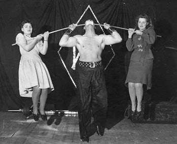 1962:  Alexander Marshall supports two women on a steel bar resting in his mouth, which is protected...
