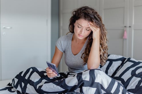 A sad young woman in bed suffers from depression and she is using a mobile phone