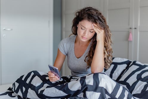 A sad young woman in bed suffers from depression and she is using a mobile phone