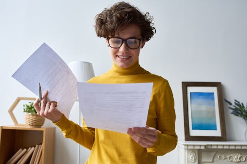 Smiling attractive curly-haired lady entrepreneur in eyeglasses standing in living room and working ...