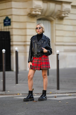 A fashion week attendee pairs a black moto jacket with a red tartan skirt and black boots. 