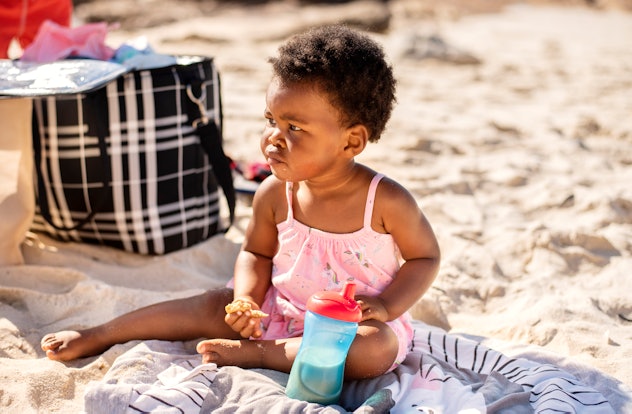 Adorable little African girl drinking from a sippy cup and eating a snack while sitting on a sandy b...