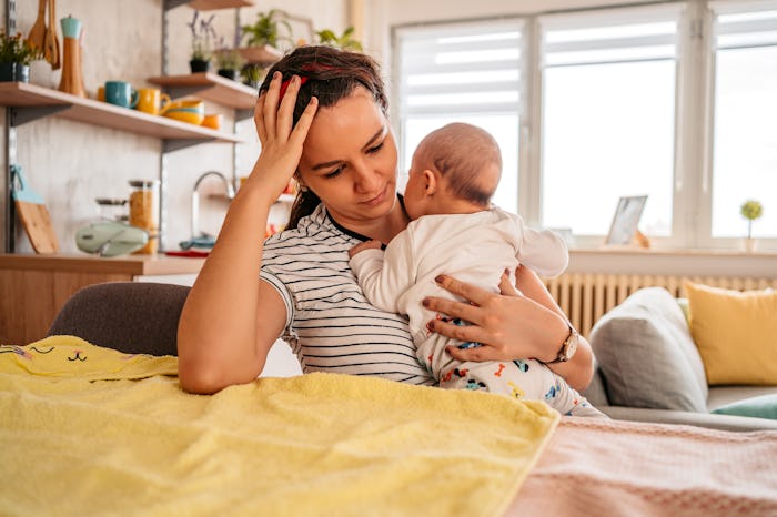 Young adult mother sitting in chair at home, holding her three month old baby son, feeling tired, sl...