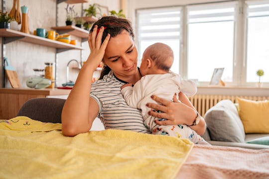 Young adult mother sitting in chair at home, holding her three month old baby son, feeling tired, sl...