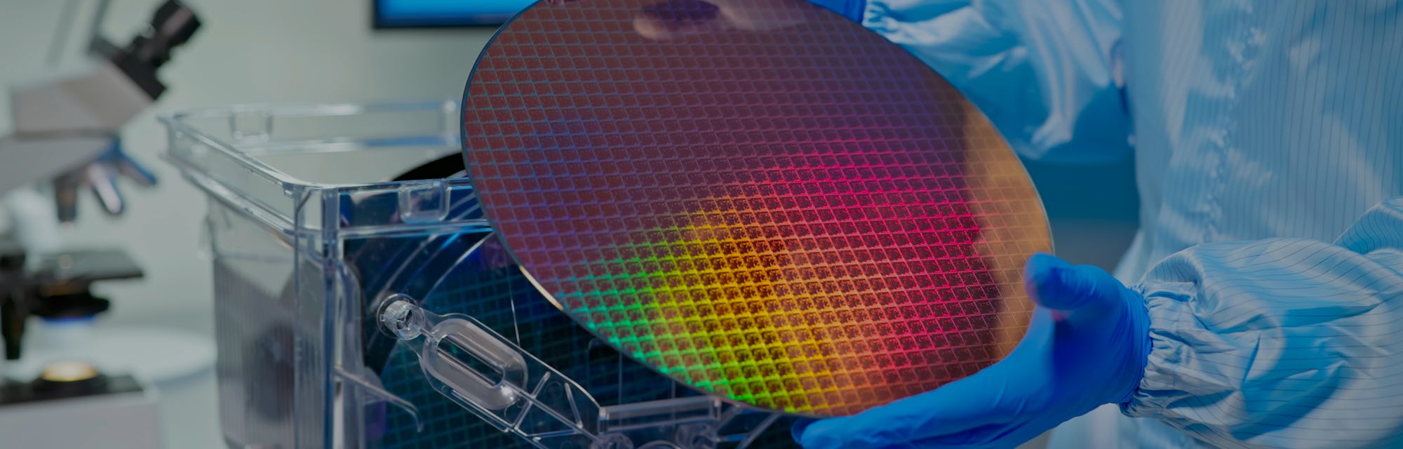 close up of asian male technician in sterile coverall holds wafer that reflects many different color...