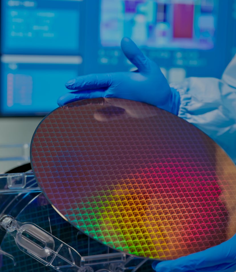 close up of asian male technician in sterile coverall holds wafer that reflects many different color...