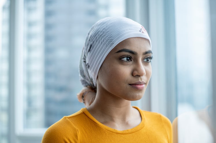 A young woman battling cancer stands in front of her apartment window gazing out with a neutral expr...