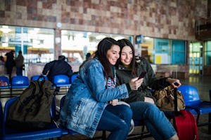 Couple of female friends looking at the smartphone while waiting for the train. Two young women chec...