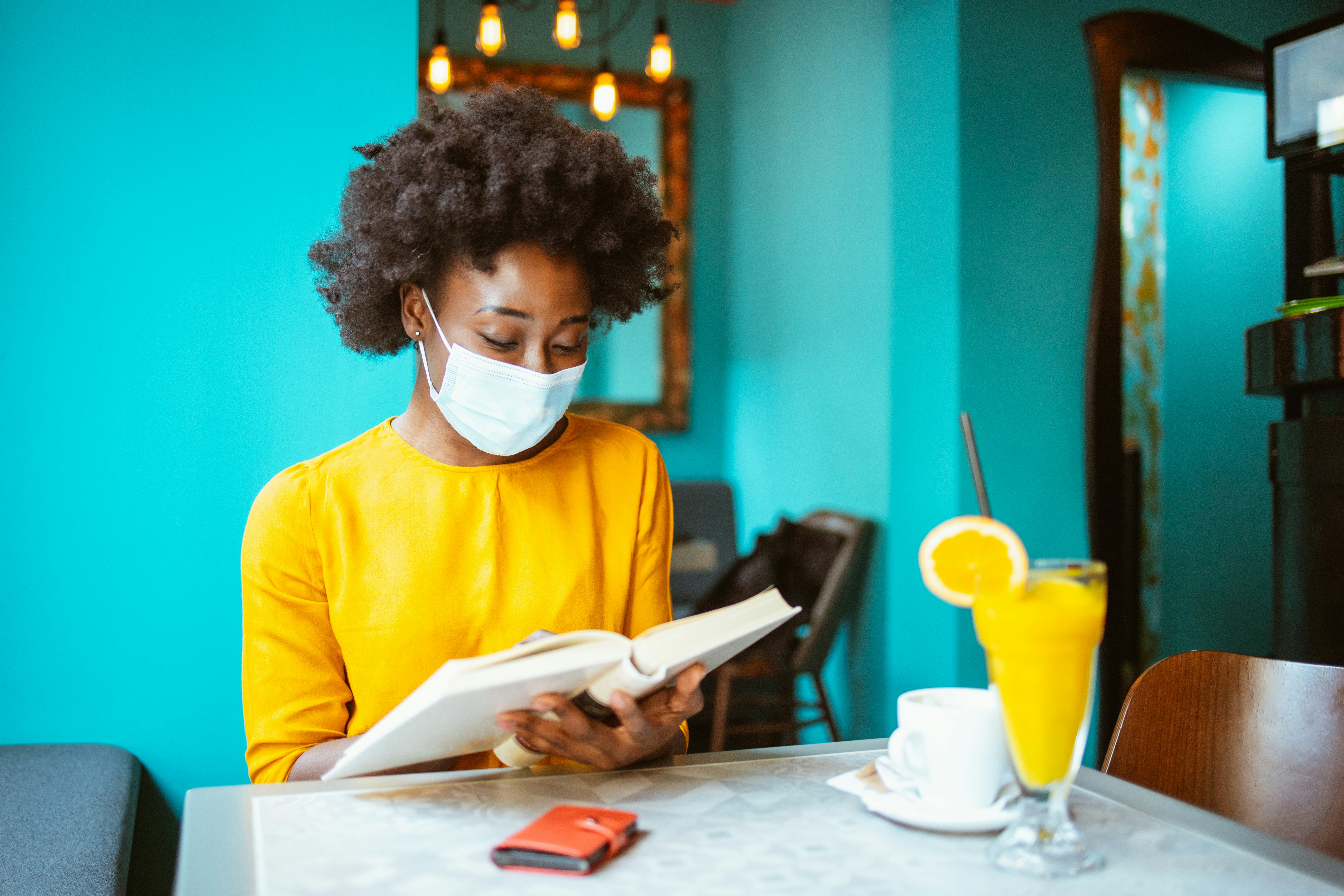 Young African-American woman enjoying reading a book and wearing protective facemask in a coffee sho...